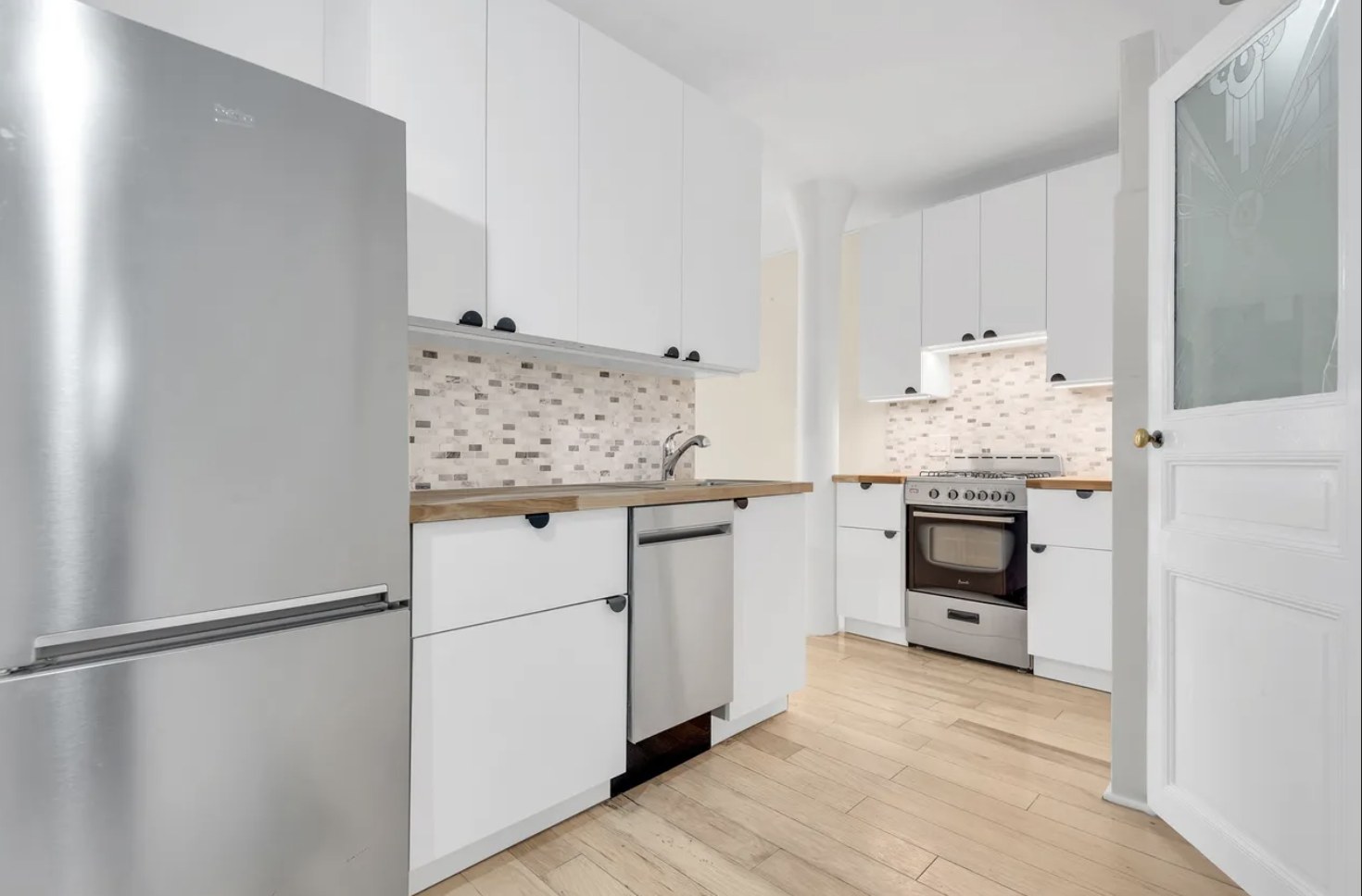 White kitchen with stainless steel appliances, light wood floors and a frosted glass door in a two-bedroom apartment on East 78th Street