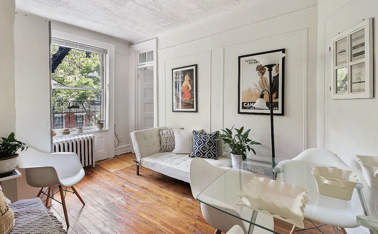 Sunlit Lenox Hill studio living area with hardwood floors, a white tufted sofa, vintage theater posters, a radiator and a tall window with a wrought-iron railing