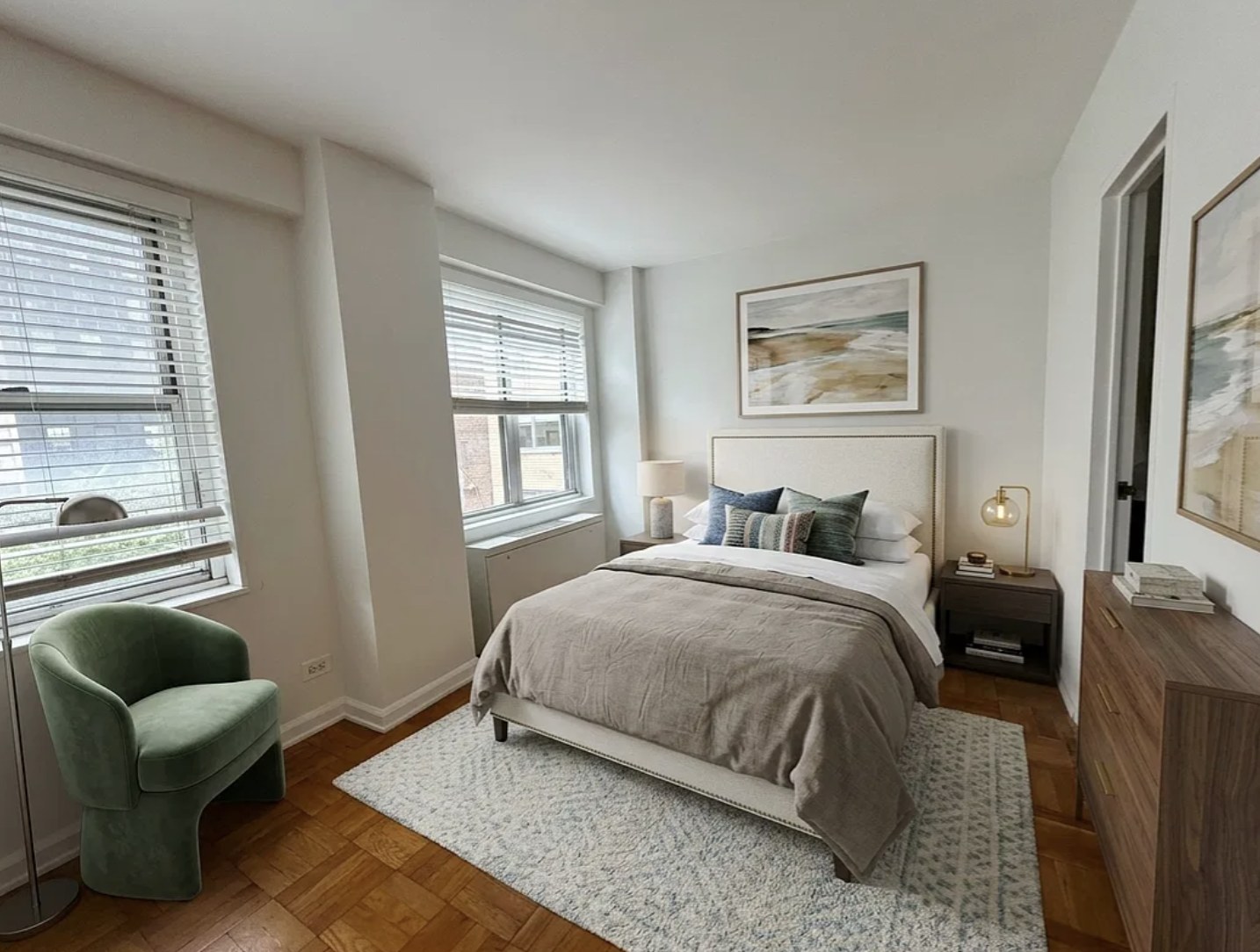 Bedroom area of a Lenox Hill studio with an upholstered bed, a green armchair, parquet floors and two windows