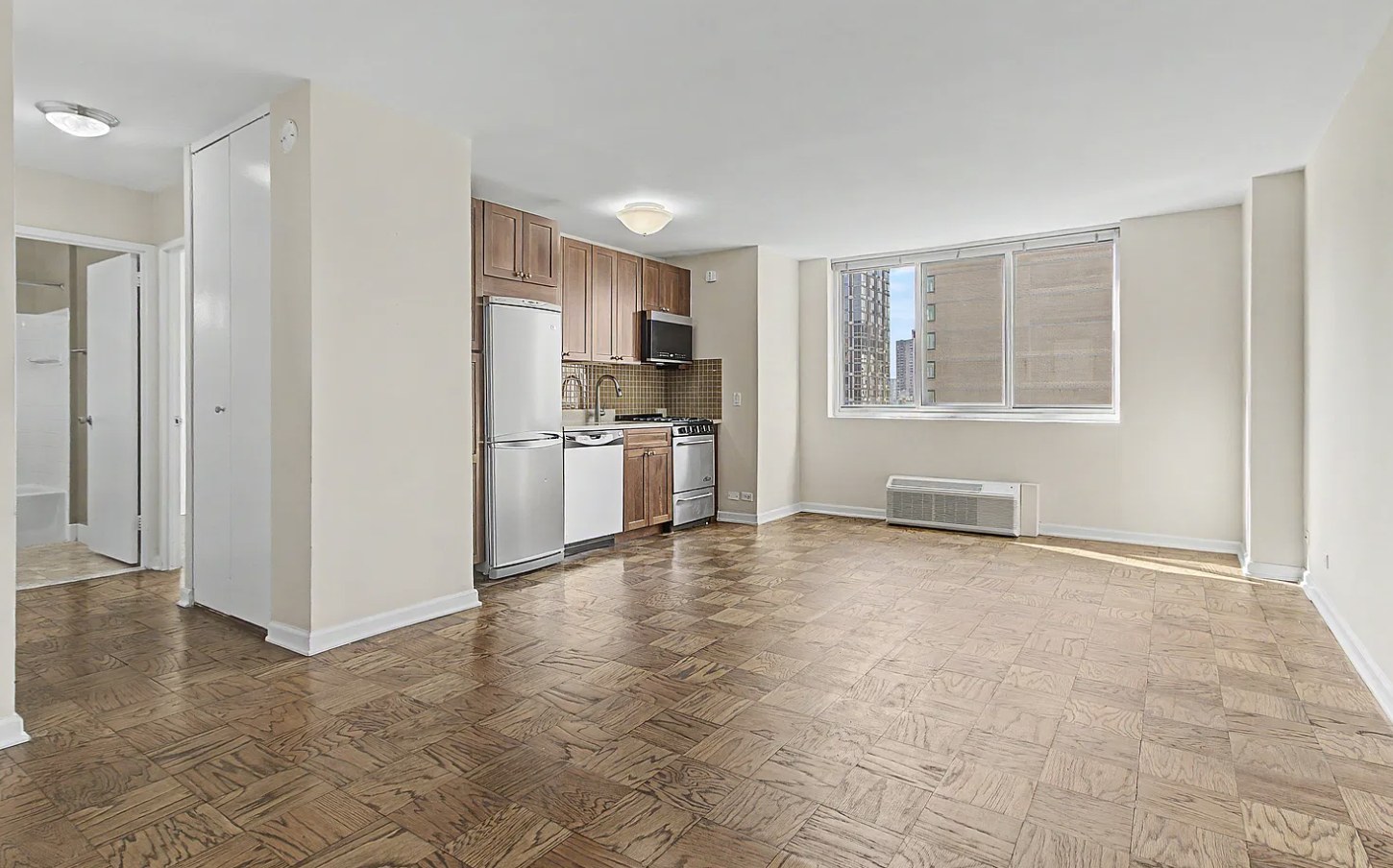Open living and kitchen area in a Yorkville one-bedroom with parquet floors, wood cabinets, stainless steel appliances and a sunny window